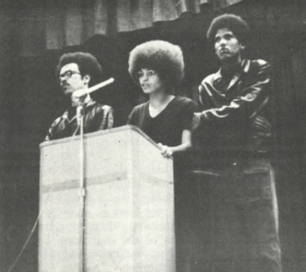 Three individuals stand behind a podium, one of them speaking into a microphone. The photo is black and white, capturing a moment of serious expression and activism.