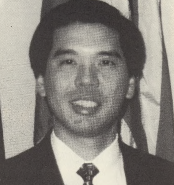 A grayscale image of a man in a suit and tie, smiling at the camera.