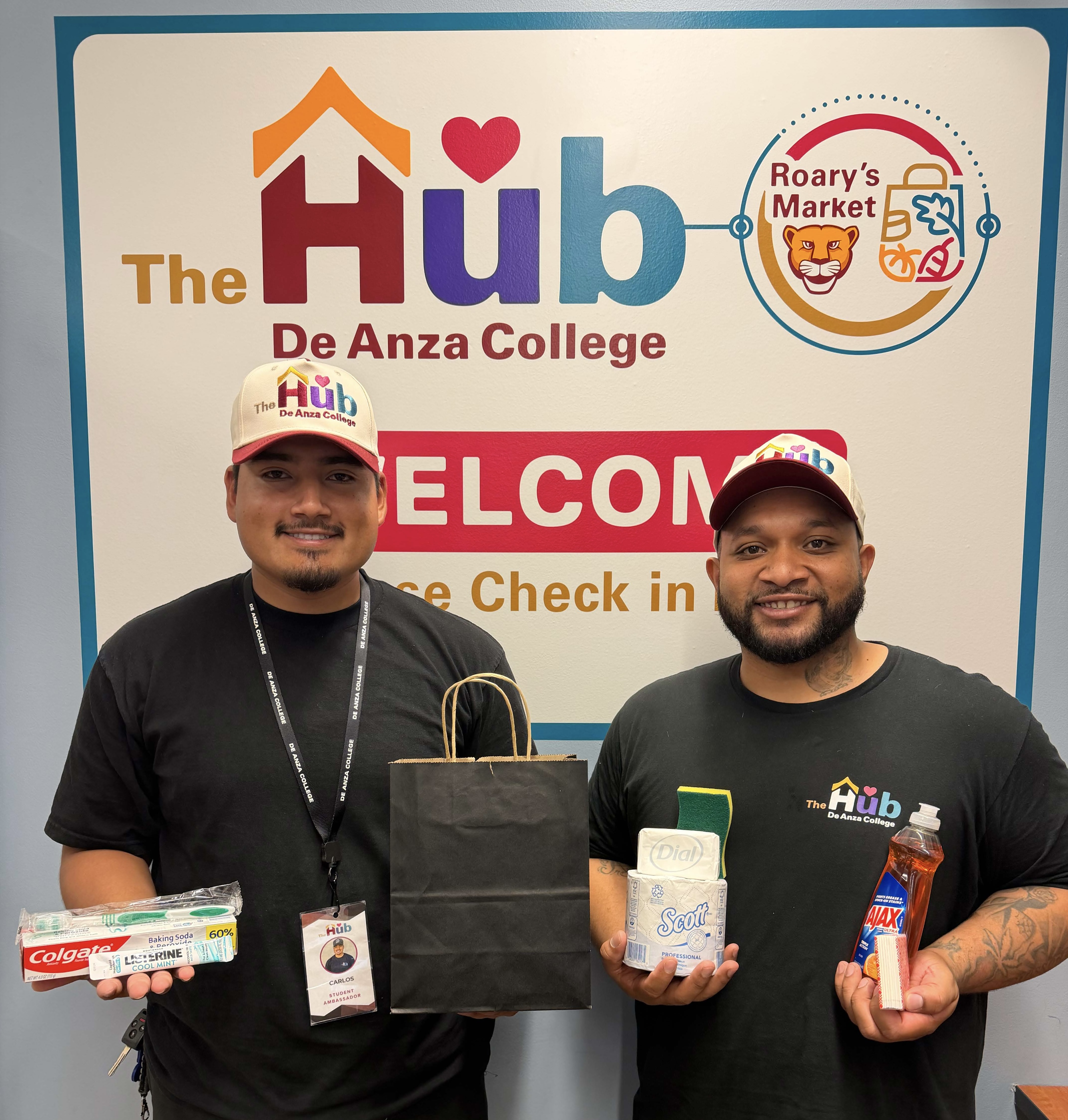 two students in black T-shirts and white baseball caps with The Hub logo on them hold supply bags and toothpaste and soap